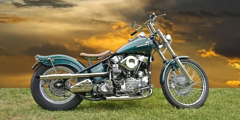 A vintage teal bobber motorcycle on grass beneath a dramatic sunset sky